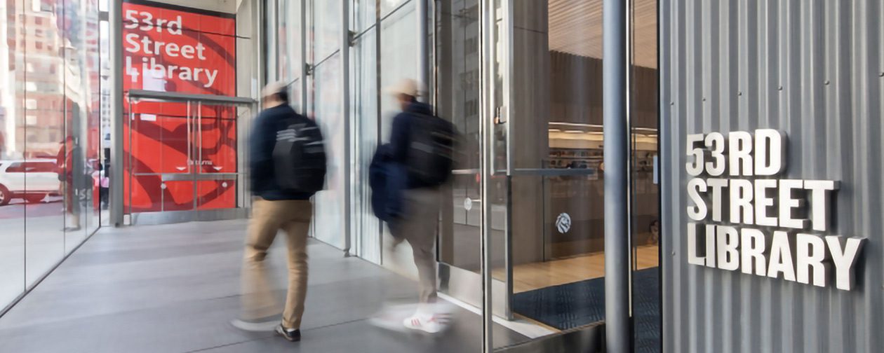 Wide-angle view of the modern glass and steel entrance to the 53rd Street Library in New York City with pedestrians walking past.