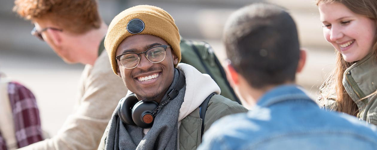 A smiling young man with glasses and a yellow beanie wearing headphones around his neck, standing outside with a group of friends.