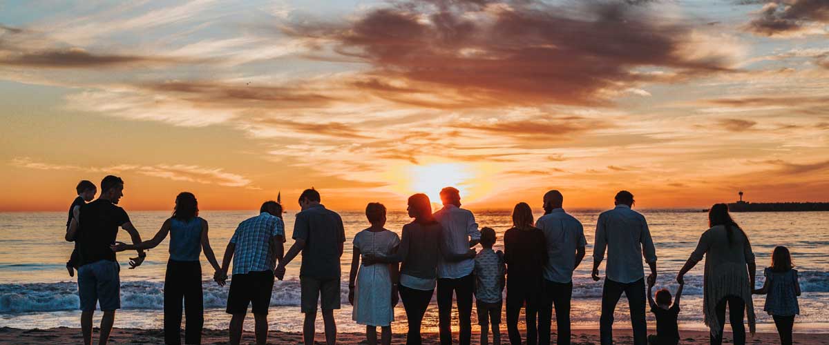 Wide panoramic silhouette of a large family holding hands on a beach during a vibrant orange sunset.