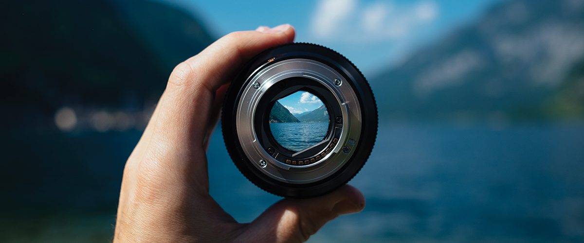 A person's hand holding a camera lens up to a scenic mountain and lake landscape, with the view perfectly centered through the glass.