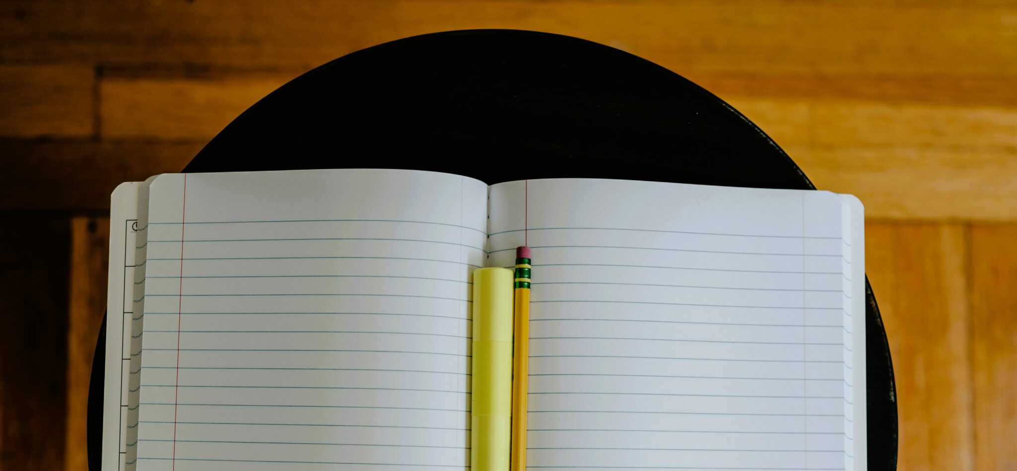 Close-up of an open, lined notebook with a yellow pencil resting in the center on a dark surface.