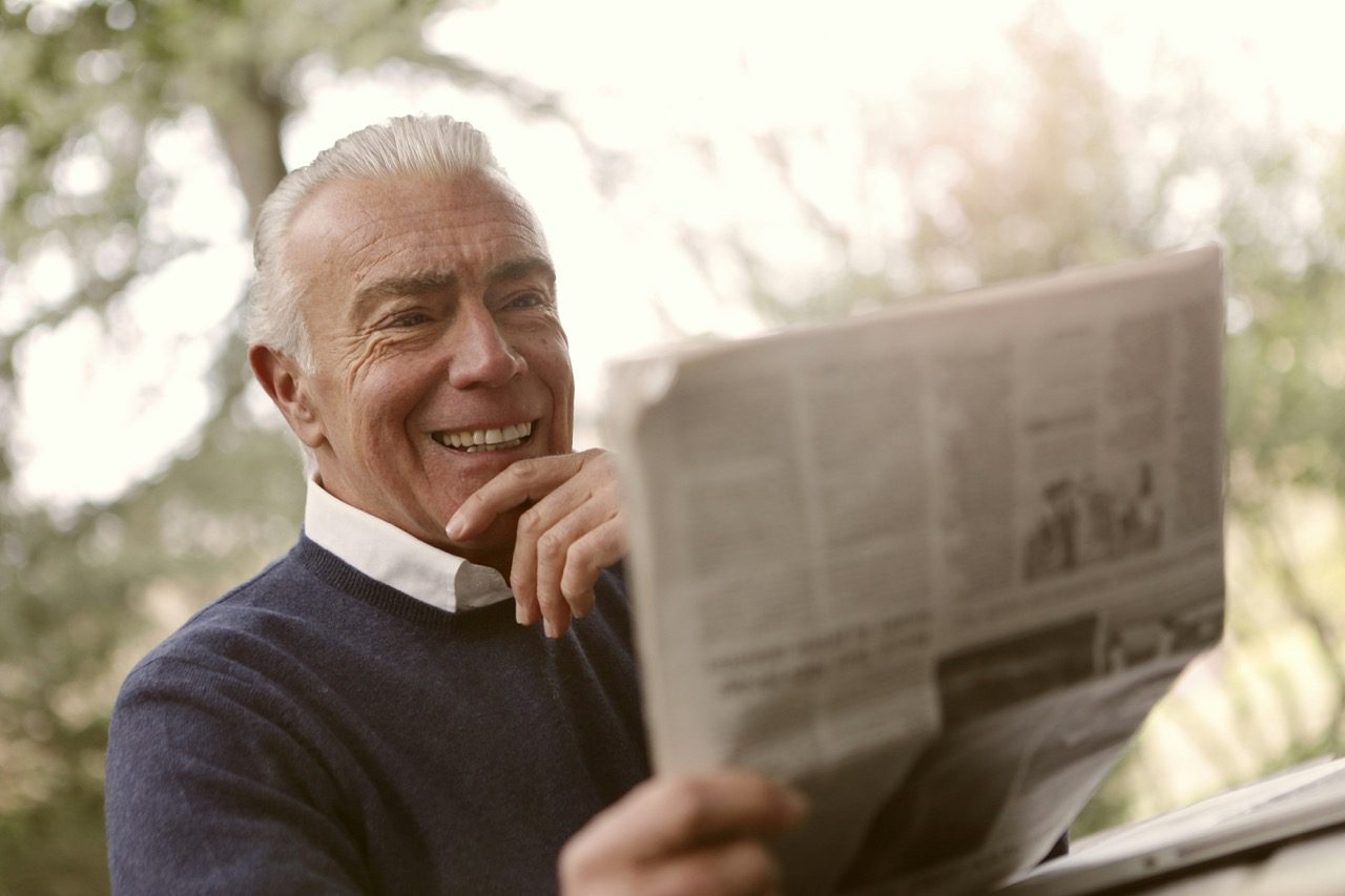 A smiling senior man with grey hair reading a newspaper outdoors in a soft-focus garden setting.