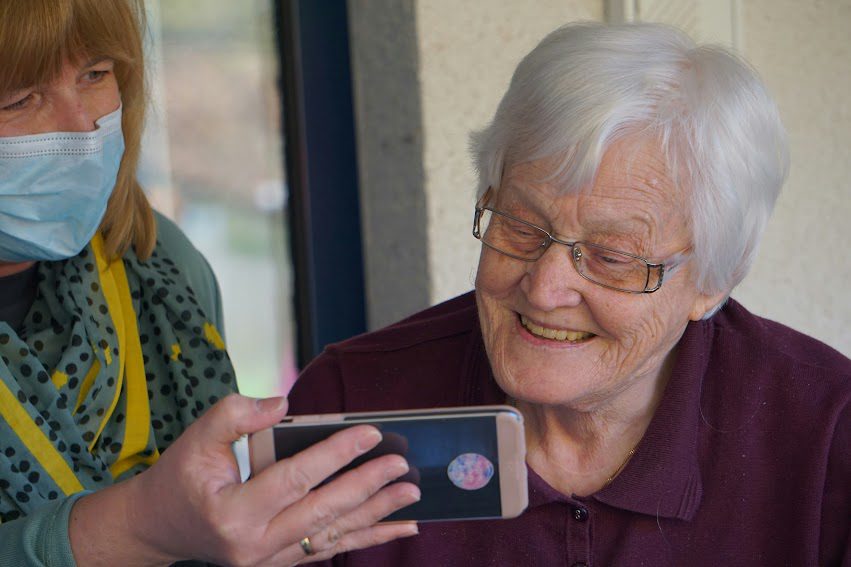 An elderly woman smiling while looking at a smartphone held by a younger woman wearing a face mask in a healthcare or retirement setting.
