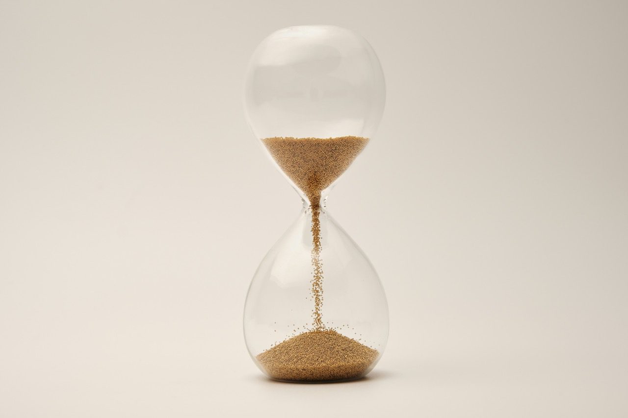 A clear glass hourglass with golden sand flowing from the top bulb to the bottom against a neutral, minimalist background.
