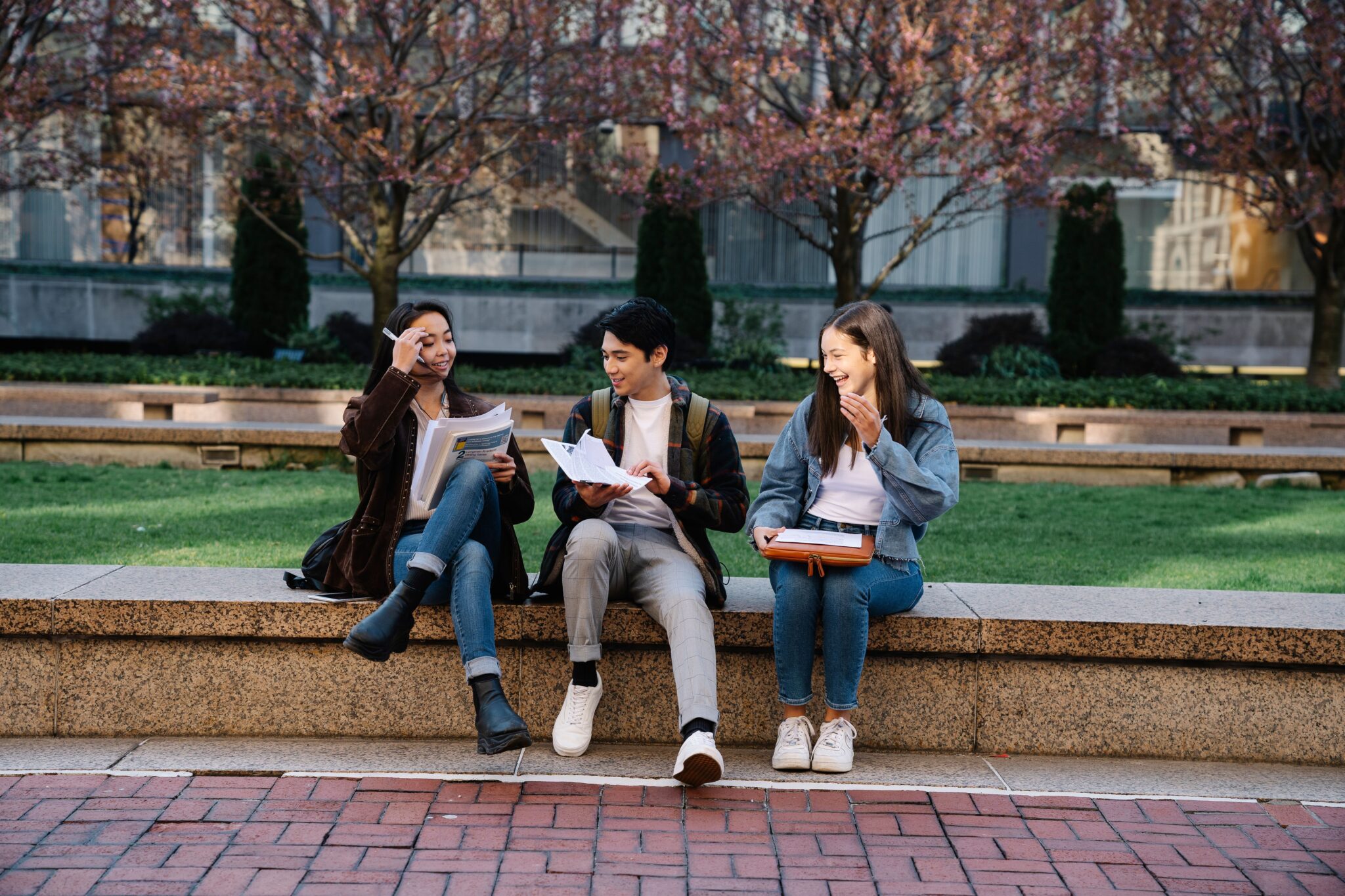 Three young adult students sitting outside on a college campus ledger, smiling and looking over study materials together.