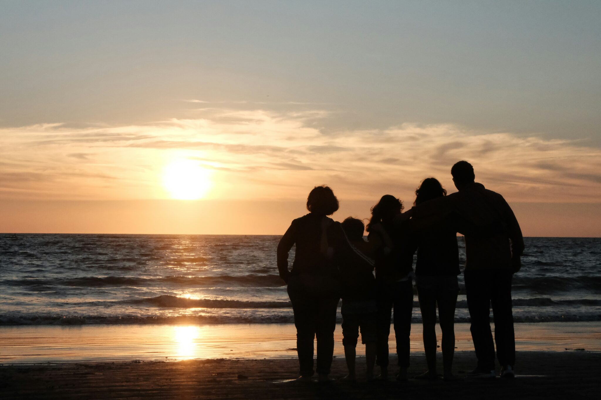 Silhouette of a family standing together on a beach facing the ocean during a golden sunset.