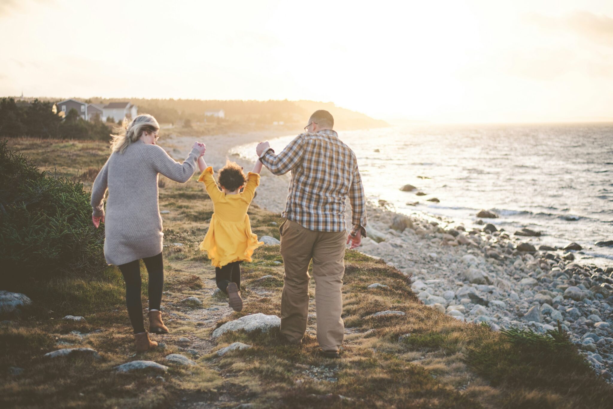 Two parents holding hands with their young child in yellow walking along a coastal path near the ocean at sunset.
